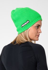 Green knitted beanie with a ribbed texture, featuring a black label with the brand "reusch" on the side. Slouchy fit and seamless design.