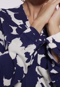 Blouse à motifs bleu marine et blanc,avec un décolleté en V, des manches longues avec des poignets boutonnés et un tissu lisse au toucher fluide.