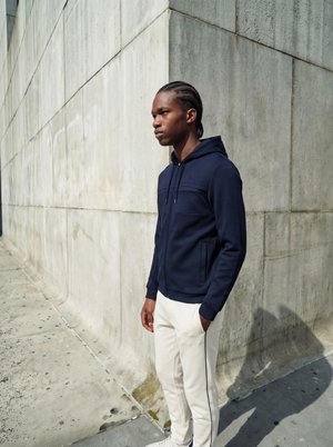 Jeune homme aux cheveux tressés se tient à côté d'un grand mur en béton gris clair, portant un sweat à capuche bleu foncé et un pantalon blanc avec des bandes latérales.