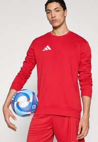 Κόκκινο φούτερ με λευκό λογότυπο Adidas, σε συνδυασμό με κόκκινο παντελόνι. Κρατάει μια μπλε και λευκή ποδοσφαιρική μπάλα με σχέδιο. Ομαλή υφή, αθλητικός σχεδιασμός.