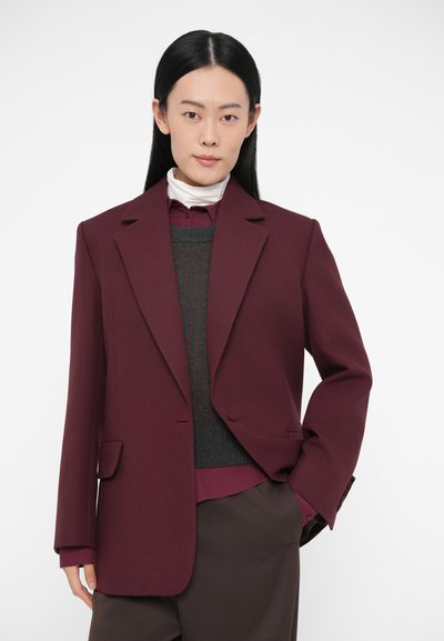 Blazer bordeaux con design strutturato, revers a lancia e tasche frontali. Indossato sopra a un maglione grigio e una camicia color granata, abbinato a pantaloni marroni.