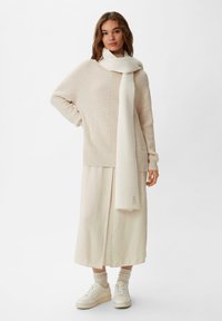 Beige gestrickter Pullover mit strukturiertem Muster, kombiniert mit einem passenden Schal, einem langen Cremé-Rock und weißen Turnschuhen. Weicher Stoff, simples Design.