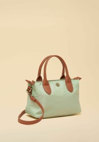 Bolso de mano verde claro con asas de cuero marrón y correa de hombro desmontable sobre fondo beige.