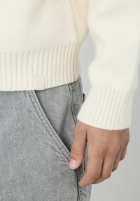 Pull en tricot couleur crème avec des poignets côtelés, associé à un pantalon en tissu gris. Gros plan sur une main reposant sur la poche.