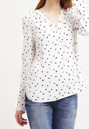 Weiße Bluse mit langen Ärmeln und schwarzem Herzmuster, V-Ausschnitt, getragen mit blauer Jeans, keine sichtbaren Accessoires oder Hintergrunddetails.