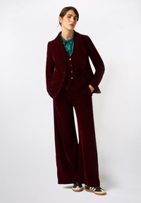 Traje de terciopelo burdeos con una chaqueta ajustada y pantalones de pierna ancha. Debajo, una camisa turquesa. Combinado con zapatillas negras y crèma.