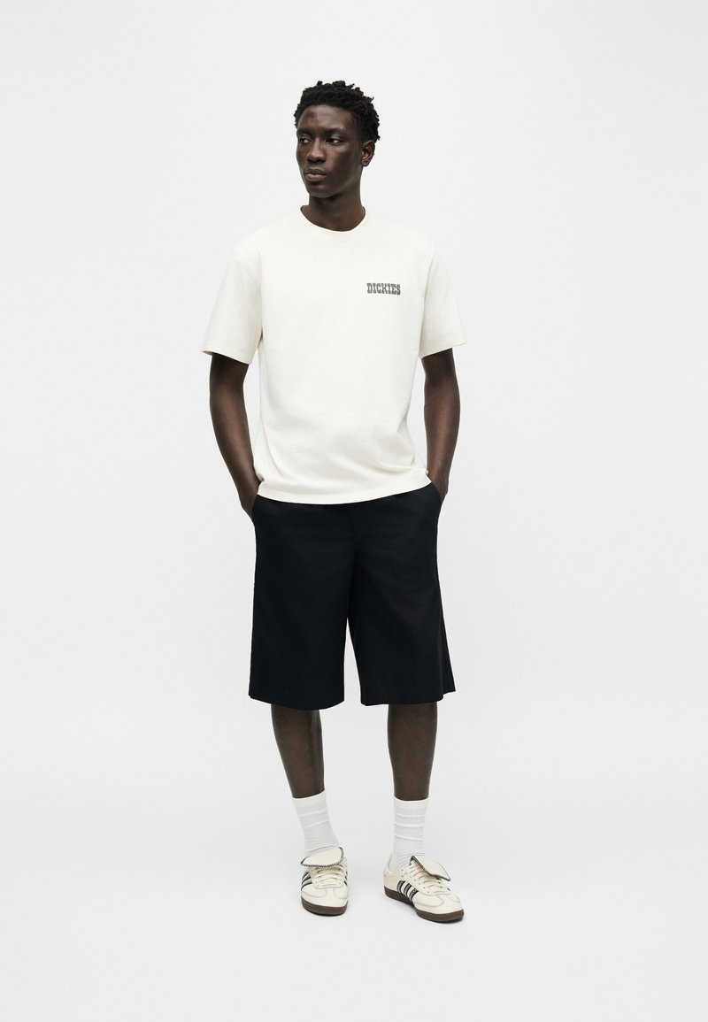 Uomo con t-shirt bianca Dickies, pantaloncini neri fino al ginocchio, calzini bianchi e sneakers beige, in piedi con le mani nelle tasche.