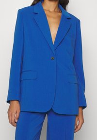 Frau trägt ein hellblaues, maßgeschneidertes Blazer und eine passende Hose, mit einem einzelnen schwarzen Knopf verschlossen und ohne sichtbares Hemd darunter.