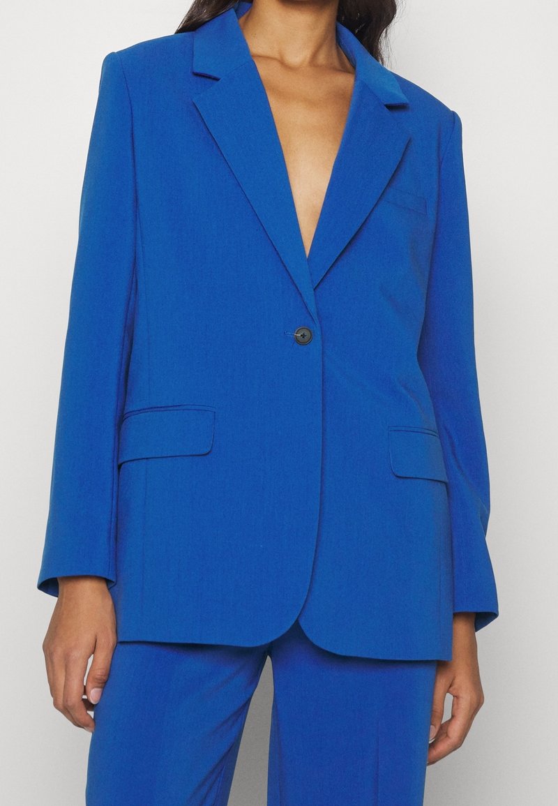 Frau trägt ein hellblaues, maßgeschneidertes Blazer und eine passende Hose, mit einem einzelnen schwarzen Knopf verschlossen und ohne sichtbares Hemd darunter.