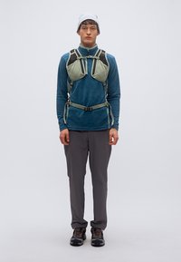 Ανοιχτό πράσινο ιμάντα με ρυθμιζόμενα λουριά, μαύρες λεπτομέρειες, φορεμένος πάνω από μπλε μακρυμάνικη μπλούζα και γκρι παντελόνι, συμπληρωμένος με καφέ παπούτσια.