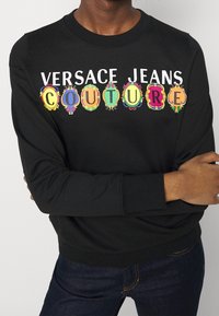 Svart sweatshirt med "VERSACE JEANS COUTURE" i fet, färgglad text, med dekorativa kanter i olika färger.