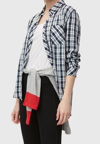 Camicia a quadri blu navy e bianca con maniche lunghe, taschino sul petto e chiusura con bottoni, abbinata a un maglione grigio legato in vita e leggings neri.