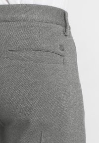 Pantalon gris texturé avec une poche horizontale à l'arrière et un accent logo en métal. Le tissu présente un motif tissé subtil.
