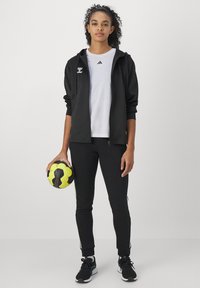 Mujer con chándal negro y camisa blanca sosteniendo un balón de balonmano amarillo y negro, de pie frente a un fondo blanco liso.
