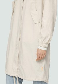 Jasno beżowy długi płaszcz wykonany z teksturowanej tkaniny. Posiada elastyczne mankiety, dwie boczne kieszenie oraz mały pomarańczowy akcent blisko dołu.