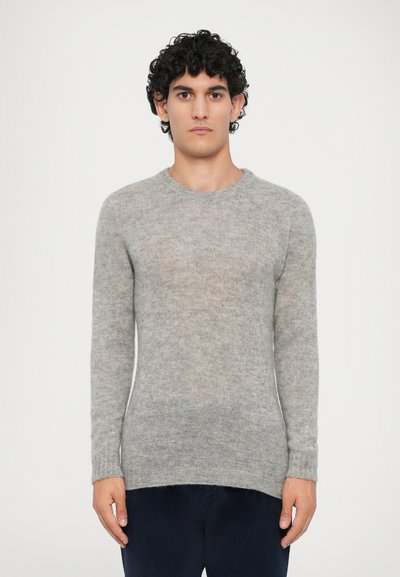 Homme aux cheveux noirs bouclés portant un pull en tricot gris clair à manches longues et un pantalon sombre, se tenant devant un fond blanc uni.