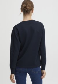 Marineblå hettegenser med lange, vide ermer og ribbestrikkede mansjetter. Mykt stoff, rund halslinje, og en avslappet passform.