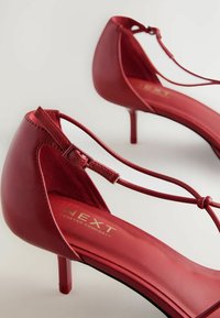 Zapatos de tacón alto rojos con un acabado de cuero elegante, correa en el tobillo, diseño de espalda abierta y punta afilada. Presentan la marca dorada "NEXT Forever Comfort".