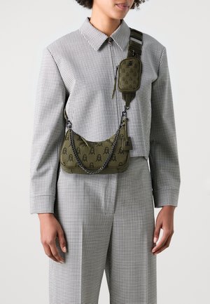 Person trägt einen schwarz-weiß karierten Anzug und zwei olivgrüne Taschen mit Kettendetails und Monogramm-Mustern quer über den Oberkörper.