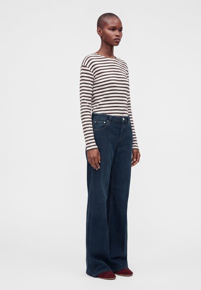 Femme debout portant un haut rayé à manches longues, un jean bleu foncé à jambes larges, et des chaussures rouges sur un fond clair uni.