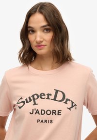 Camiseta de algodón de color rosa claro con el texto impreso en negro "SuperDry J'ADORE PARIS" en un diseño curvado, con mangas cortas y cuello redondo.