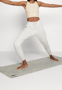 Top corto beige e pantaloni della tuta bianchi con cordoncino, aderenti alle caviglie, su un tappetino da yoga testurizzato. La modella sta eseguendo una posa di yoga.