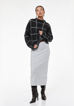 Vrouw die een zwart-grijs geruite trui draagt, een lichtgrijze kokerrok, zwarte laarzen met hakken en oorringen tegen een witte achtergrond.