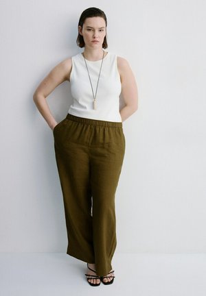 Kobieta stojąca przy białej ścianie, ubrana w biały, bezrękawnik, oliwkowe spodnie z szerokimi nogawkami, czarne sandały oraz długi naszyjnik z zawieszką.