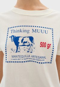 Witte katoenen t-shirt met een blauwe afbeelding van een koe, gelabeld "Thinking MUUU" en "500 gr," met blauwe gestreepte randen en rode tekstdetails.