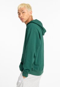 Felpa con cappuccio verde in tessuto morbido, con tasca a marsupio e polsini a coste. Indossata con pantaloni della tuta grigi. Design semplice e funzionale.