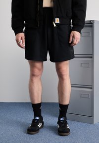 Black zip-up bomber jacket, black textured shorts, black Adidas sneakers with white stripes, and black socks. Gray filing cabinet in background.