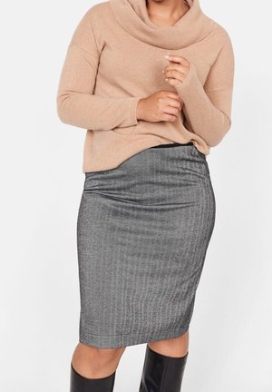 Femme portant un pull beige à manches longues, une jupe crayon grise mi-genou et des bottes noires montantes, sur fond uni.