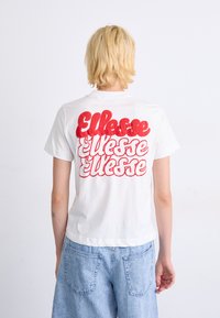 Hvit bomull T-skjorte med stor rød retro-stil "Ellesse" tekst på ryggen. Korte ermer, rund halsutringning og avslappet passform, kombinert med denimshorts.