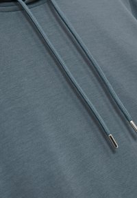 Felpa blu con una texture liscia, caratterizzata da un laccetto in contrasto e punte metalliche, che evidenziano un design rilassato e una finitura semplice.