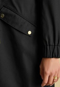 Čierny kabát s klopovým vreckom, mosadznými patentkami a embossovaným textom "JOULES", s naťahovacími manžetami a hladkým textúrou látky.