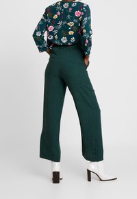 Gröna vid byxor i strukturerad tyg, tillsammans med en topp med blommönster. Vita klackstövlar fulländar outfiten.