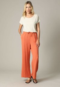 Lichtgewicht crèmekleurig topje met korte mouwen; losse, hooggesneden oranje broek met elastische tailleband en strikdetail; platte sandalen.