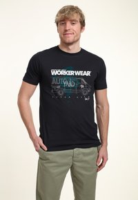 Černé bavlněné tričko s grafikou "WORKER WEAR" a ilustrací dodávky se textem, spárované s olivově zelenými kalhotami.