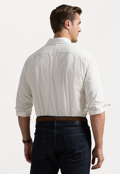 Λευκό πουκάμισο με ρολαρισμένα μανίκια, φορεμένο με σκούρα τζιν και καστανή δερμάτινη ζώνη. Απλός σχεδιασμός, ελαφρύ ύφασμα.