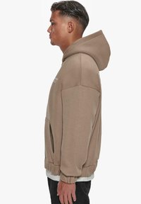 Beige hooded sweatshirt with ribbed cuffs, front kangaroo pocket, and soft texture. Features a logo printed on the chest.