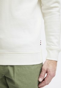Felpa bianca in tessuto testurizzato con un piccolo logo a righe in evidenza all'orlo. I polsini sono aderenti e viene abbinata a pantaloni verdi.