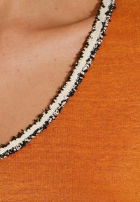 Oranje textiel met een zachte textuur heeft een V-halslijn afgewerkt met een contrasterende witte en zwarte gestikte rand.