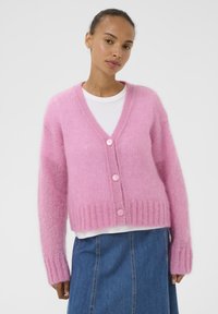 Rosa, flauschige Strickjacke mit V-Ausschnitt, geripptem Saum und drei Knöpfen vorne. Getragen über einem weißen Hemd, kombiniert mit einem Jeansrock. Weiche Textur.