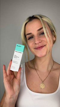 Kvinde med blondt hår holder en æske med Dr.Jart+ Pore Remedy PHA Exfoliating Serum, smilende, iført hvidt tanktop og guldvedhængshalskæde.
