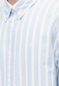 Ανοιχτό μπλε και λευκό ριγέ πουκάμισο με κουμπιά, κατασκευασμένο από υφασμάτινη ύλη με υφή, με μικρό κεντητό λογότυπο στο στήθος και κανονικό γιακά.