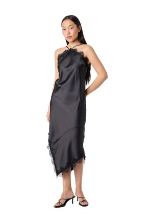 Nainen mustassa hihattomassa satiinimekossa, jossa on pitsireunus ja epäsymmetrinen helma, mustat remmilenkkikorkokengät jalassaan, seisoo valkoisella taustalla.