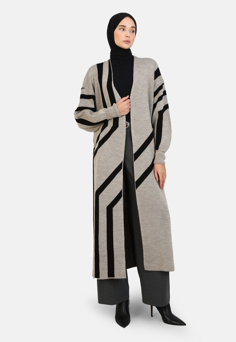 Long, open-front cardigan in beige with bold black diagonal stripes. Knit fabric, loose fit, and ribbed cuffs. Paired with dark trousers and heels.