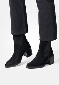 Botines negros de ante con tacón cuadrado, paneles laterales elásticos y textura suave. Posicionados con jeans negros, mostrando su diseño.