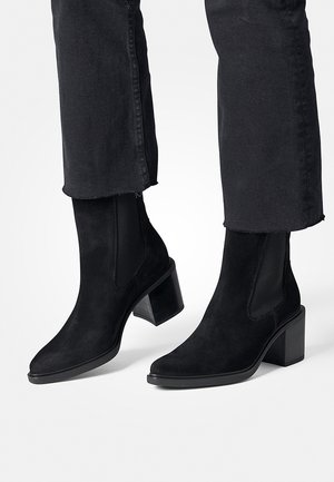 Stivaletti neri alla caviglia in suede con tacco quadrato, pannelli laterali elastici e una texture liscia. Posizionati con jeans neri, che mettono in risalto il loro design.