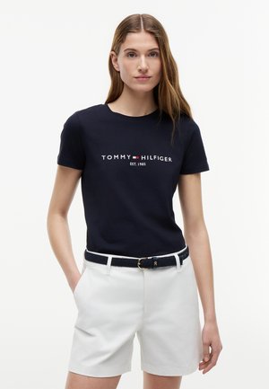 Frau trägt ein marineblaues Tommy Hilfiger T-Shirt und weiße Shorts mit einem navyfarbenen Gürtel, steht mit einer Hand in der Tasche vor einem schlichten Hintergrund.